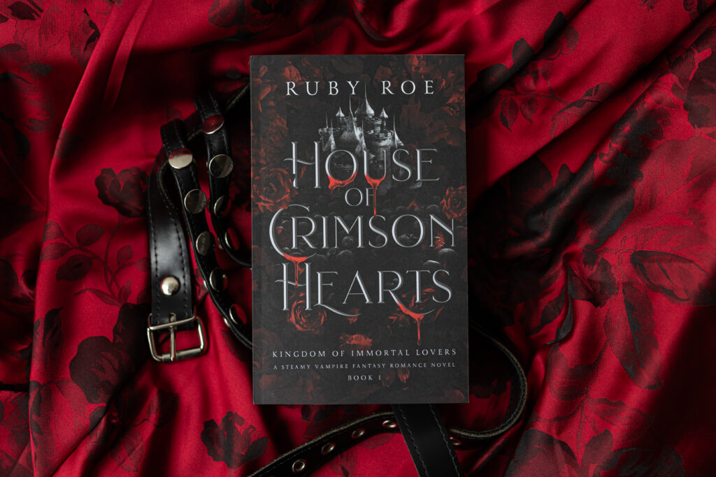 Image of the paperback book surrounded by lush red blanket and black studded belt decoration