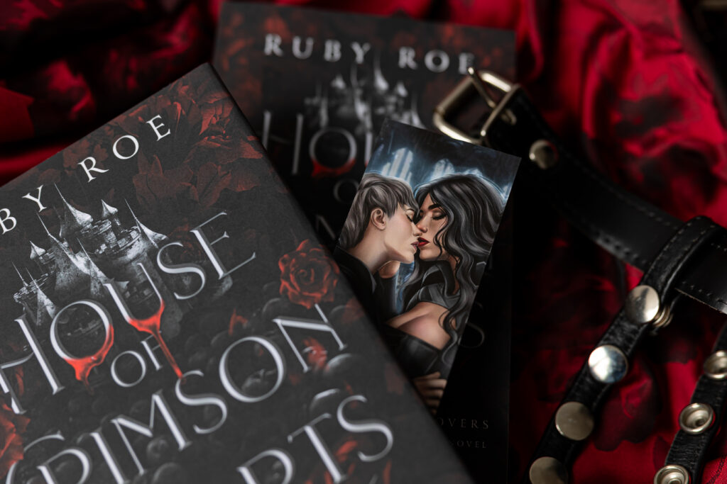 Image of the hardback with a bookmark of the two love interest kissing, surrounded by a blanket of red fabric and black belt decoration