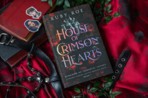 Image of the book: House of Crimson Hearts on a red silky blanket with decorative pieces around it including character stickers and green foliage.