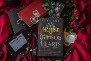 Image of the book: House of Crimson Hearts laid on a red silky blanket with some greenery and decorative items including character stickers and certificate of authenticity